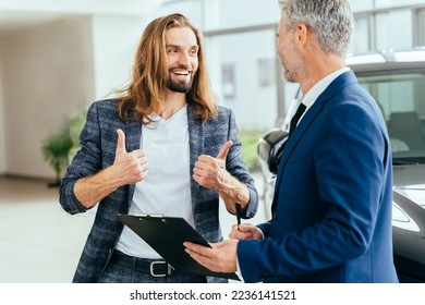 Young buyer happy with car rental terms showing thumbs up. Successful middle-aged businessman in a business suit is selling cars to a young hansome male buyer in a car showroom. - Shutterstock ID 2236141521