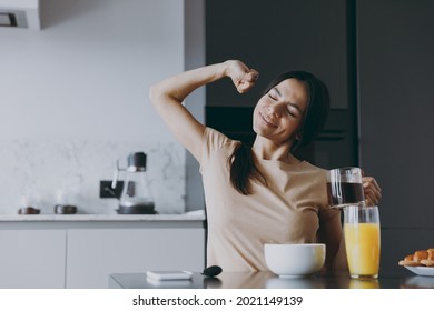 Young calm awaken housewife woman 20s in casual beige t-shirt eat breakfast hold cup of tea drink coffee do stretch morning cook food in light kitchen at home alone. Healthy diet lifestyle concept. - Powered by Shutterstock