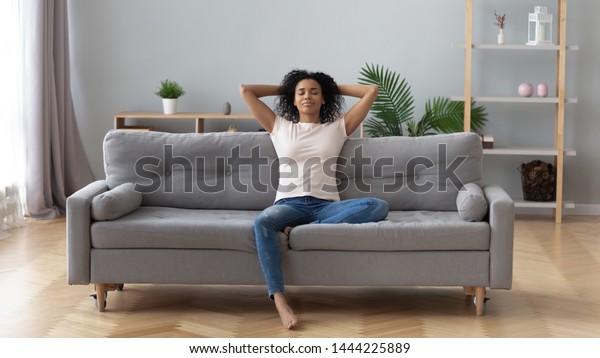 Young calm black woman relaxing sit on comfortable\
sofa in modern living room, lazy happy african woman girl resting\
on couch breathing fresh air enjoy peace of mind no stress free on\
couch at home