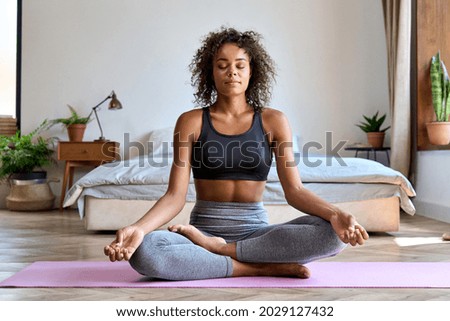 Young calm fit healthy African American woman wearing sportswear sitting at home in bedroom doing yoga exercise, meditating and breathing in the morning. Mental health and zen meditation concept