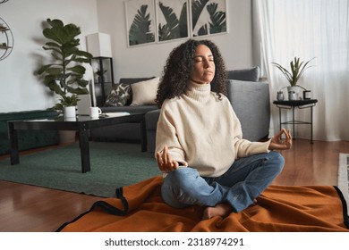 Young calm latin woman doing yoga exercise at home feeling zen. Mindful girl sitting on floor meditating chilling with eyes closed, relaxing breathing for good mental balance, peace of mind.