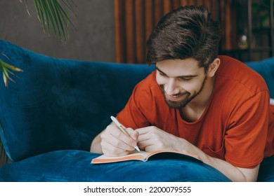 Young Calm Man Wear Red T-shirt Writing Memories In Notebook Diary Lay Down On Blue Sofa Stay At Home Hotel Flat Rest Relax Spend Free Spare Time In Living Room Indoors Grey Wall People Lounge Concept