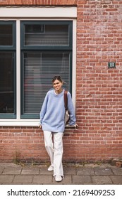 Young Calm Smiling Freelance Woman In Blue Sweater And White Pants Looking At Camera, Standing Outdoors On Red Brick Building House, Big Window Background. Portrait Of Happy Blonde Female With Low Bun