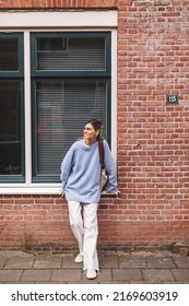 Young Calm Smiling Freelance Woman In Blue Sweater And White Pants Looking At Side, Standing Outdoors On Red Brick Building House, Big Window Background. Portrait Of Happy Blonde Female With Low Bun.