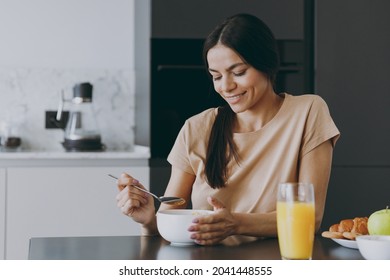 Young Calm Smiling Happy Brunette Housewife Caucasian Woman 20s Wear Casual Clothes Beige Shirt Eat Oatmeal Porridge Muesli Cooking Food In Light Kitchen At Home Alone. Healthy Diet Lifestyle Concept