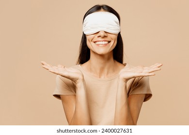 Young calm smiling happy Latin woman wear pyjamas jam sleep eye mask rest relax at home cover eyes spread arms hands isolated on plain pastel light beige background studio. Good mood night nap concept