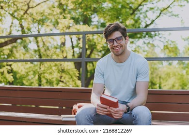 Young Calm Smiling Happy Student Man 20s In Blue T-shirt Eyeglasses Read Book Sitting On Bench Walking Rest Relax In Sunshine Spring Green City Park Outdoors On Nature. Education High School Concept