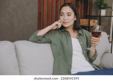 Young calm woman of Asian ethnicity wear casual clothes drink red wine look aside in pensive mood sits on grey sofa couch stay at home hotel flat rest relax spend free spare time in living room indoor - Shutterstock ID 2268541595