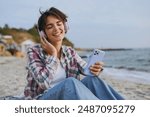 Young calm woman wear shirt casual clothes headphones sits listen music use mobile cell phone listen music rest on sea ocean sand shore beach outdoor seaside in summer day free time. Lifestyle concept