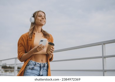 Young calm woman wearing orange shirt beige top casual clothes listen to music in headphones use mobile cell phone drink coffee rest relax walk outside in summer day. Urban lifestyle leisure concept - Powered by Shutterstock