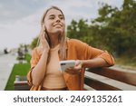 Young calm woman wears orange shirt casual clothes use mobile cell phone listen to music in earphones sit on bench walk rest relax in spring green city park outdoors on nature. Urban lifestyle concept