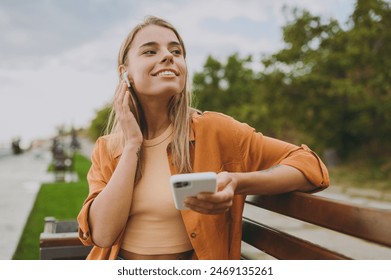 Young calm woman wears orange shirt casual clothes use mobile cell phone listen to music in earphones sit on bench walk rest relax in spring green city park outdoors on nature. Urban lifestyle concept