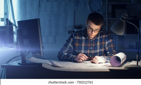 Young Capable Architectural Engineer Working at Night in His Office. Office is Dark Only His Table Light is On. Various Plans and Blueprints Lying on the Table. He Draws in His Notepad.