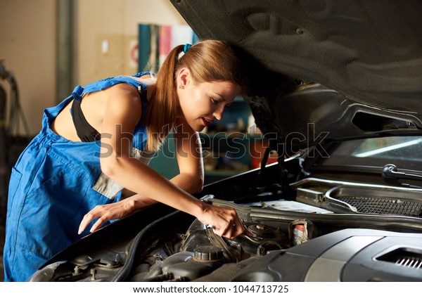 Young car mechanic woman repairing car at service\
station. Bautiful woman with wrench in her hand, dressed in blue\
work uniform