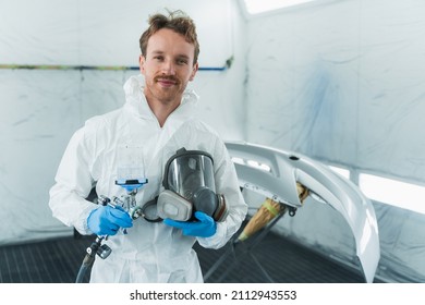 Young Car Paint Service Worker Stands In A Automobile Painting Booth With A Gun And A Protective Mask In His Hands. 
Portrait Of A Car Painter