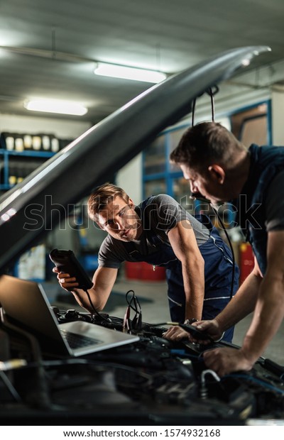 Young car repairman talking with his coworker while\
checking engine performance with diagnostic tools in auto repair\
shop. 
