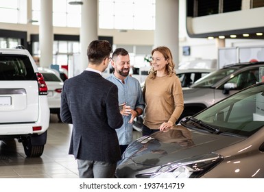 Young Car Salesman Showing To Mid-age Couple New Automobile At Dealership Salon.