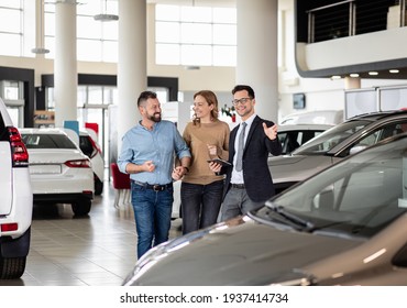 Young Car Salesman Showing To Mid-age Couple New Automobile At Dealership Salon.