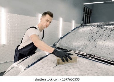 Young Car Service Male Worker In Black Rubber Gloves Doing Cleaning The Hood Of The Car With The Help Of Shampoo And Yellow Sponge In Car Washing Company