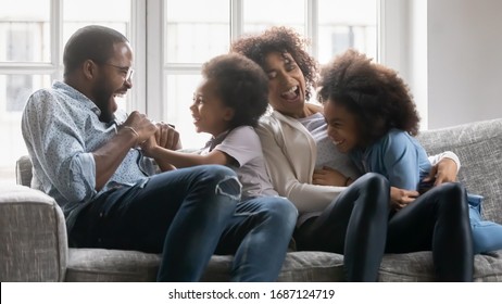 Young Carefree African Ethnicity Couple Play With Little Daughter And Son, Full Family Seated On Sofa In Living Room Tickling Each Other Laughing Feels Happy Enjoy Free Time Weekend Activities At Home