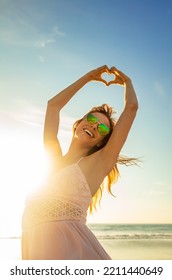 Young Carefree Bohemian Girl Enjoying Blue Sky And Summer Sun Dancing On Beach Showing Heart Sign With Hands 