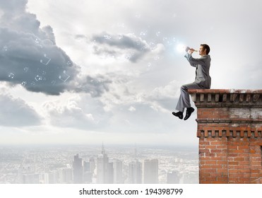 Young Carefree Businessman Sitting On Top Of Building And Playing Flute