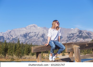 Young Carefree Smiling Woman With Windblown Dark Blond Hair With Highlights In Casual Outfit Sitting On Bridge Rail Looking Away Enjoying Summer Sunny Windy Day In Banff National Park, Alberta, Canada