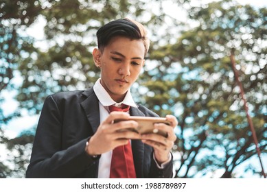 A young carefree student plays an engaging online MOBA game on his cellphone while walking around a park. Disheveled college uniform.
