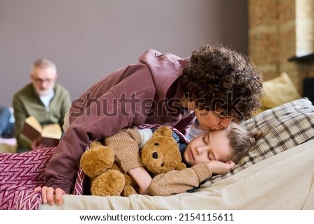 Young careful mother giving her adorable sleeping son good night kiss while bending over him against homeless mature man
