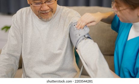 Young Caregiver Doctor Examining Senior Patient Using Blood Pressure Gauge, Caregiver Nurse Taking Care Of Elderly Grandfather Sitting On Sofa At Home Visit, Medical Service Concept