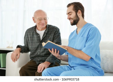 Young Caregiver Reading Book To Senior Man At Home