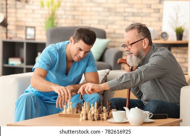 Young Caregiver And Senior Man Playing Chess At Home