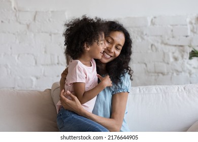 Young caring African mother sit on sofa hugging cheerful laughing little 5s daughter, loving family enjoy time together, embracing feeling unconditional love. Happy motherhood, bond, custody concept
