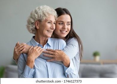 Young caring grand daughter hugging to loving elderly grandmother, multi-generational relatives women standing look in window dreaming feels happy spend time together, love connection warmth concept