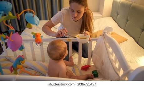 Young Caring Mother Feeding Her Baby Son With Soup In Baby Crib At Night. Concept Of Healthy Nutrition, Parenting And Baby Care.