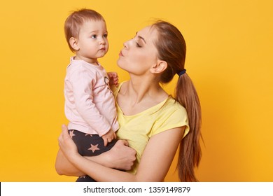 A Young Caring Mother Tries To Calm Down Her Crying Daughter. A Tear Is Going To Run Down Cute Baby`s Cheek. A Disappointed Child Looks Concentrated On Something Aside. Mum Kisses Her Amazing Kid.