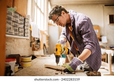 Young carpenter drilling hole in wood with drill at workshop