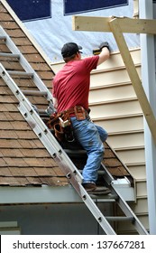 Young Carpenter Remodels Aging Home.  He Is Installing Siding And Is Using A Level To Be Sure Installation Is Precise.  He Is Standing On A Ladder On Rooftop.