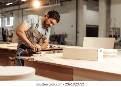 Young carpenter sanding wood piece in workshop in furniture factory - Shutterstock ID 2361923721