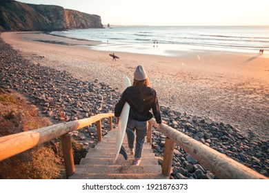 Young casual dressed woman walk down wooden stairs or walk board to epic beach at sunset, carry professional surfboard ready for evening surf session. Millennial hobby or surfing lifestyle - Powered by Shutterstock