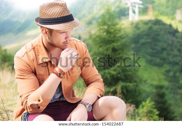 young casual man sitting in\
the nature with a straw in his mouth and looking away from the\
camera