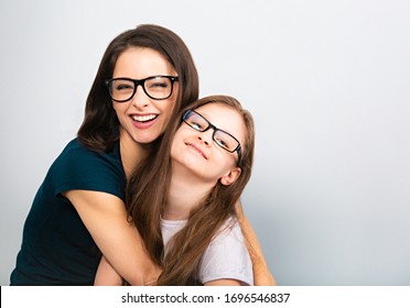 Young Casual Toothy Smiling Mother And Happy Kid In Glasses Hugging On Light Blue Background. Closeup Studio Portrait. Teaching Kids Staying At Home. Online Education. Distance Learning