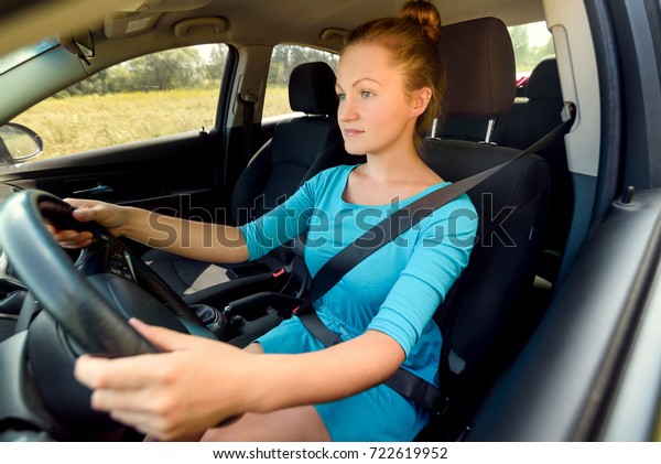 Young casual woman in blue dress driving a car, side\
view. Beautiful young girl at the wheel of car with black interior\
looking at the road