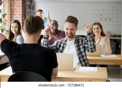 Young casually dressed male office worker throws hands in air celebrating achievement at work. Coworkers around cheer and clap hands. Rewarding out come, received promotion, achieved success concept. 