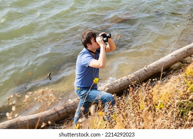 Young Caucasian 34 Years Old Man Is Looking Through Binoculars On Lake Coast. Summer Hiking, Leisure Activities, Bird And Animal Watching On River. Lifestyle