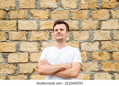 Young Caucasian 34 Years Old Man In White T-shirt With Cross Arms Chest On Stone Bricks From Limestone Laid Background. Waist Up Lifestyle Looking Away Unshaven Man Portrait. Entrepreneur, Businessman