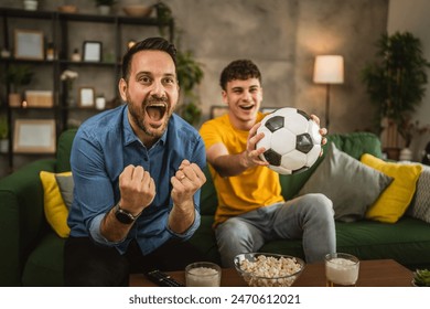 Young caucasian and adult caucasian man brothers or friends watch football and cheer match hold football ball with popcorn and beer on table at home - Powered by Shutterstock