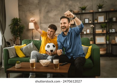 Young caucasian and adult caucasian man brothers or friends watch football and cheer match hold football ball with popcorn and beer on table at home - Powered by Shutterstock