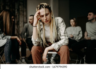 Young Caucasian Alcoholic Woman Tired Of Conviction And Gossip, Sit In The Center Of Room While People In The Background Discuss Problems Connected With Alcohol. Support, Problems, Help, Desperate