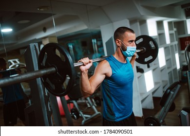 A Young Caucasian Athlete Man With A Mask On His Face Exercises And Lifts Weights In The Gym. COVID 19 Coronavirus Protection